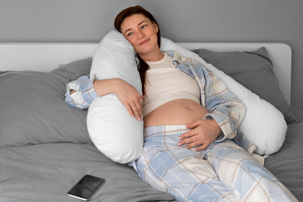  Pregnant woman in plaid pajamas relaxing on a bed with a white pregnancy pillow, gently cradling her belly and smiling peacefully. A smartphone is placed on the bed beside her.