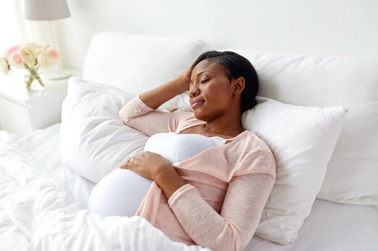 Pregnant woman sleeping comfortably in bed at home, wearing a light pink top and holding her belly, with white bedding and a vase of flowers in the background.